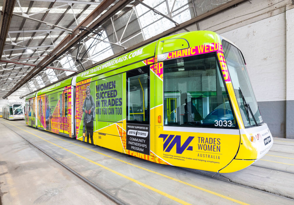 Photo of a C1 tram in a maintenance shed, wrapped in a yellow, green and pink design from Tradeswomen Australia, including the wording