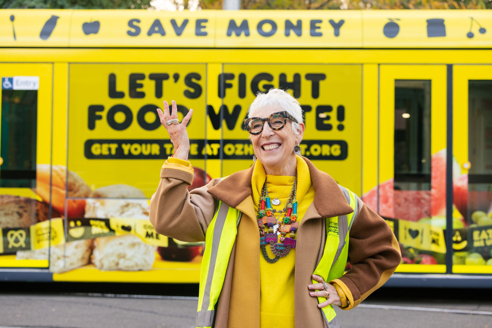 Photo: Ronni Kahn AO, OzHarvest CEO and Founder poses in front of a vibrant yellow tram, which has images of food and the text 