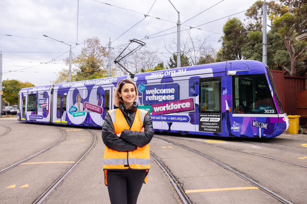Woman in an orange vest stands in front of a purple tram with large text spelling out 